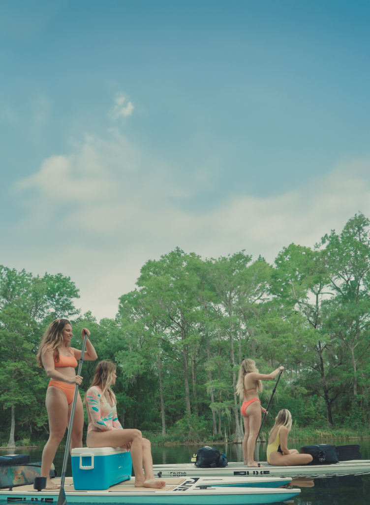 This image showcases two pairs of adventurous girls confidently paddling on LIVE Watersports Paddle Boards in Florida waters. Seated on each board is a companion, gearing up for a dive with the innovative Nomad Dive Systems comfortably packed in backpacks on the boards. This picture perfectly encapsulates the spirit of outdoor adventure, friendship, and the excitement of diving made accessible and simple through Nomad Dive Systems.