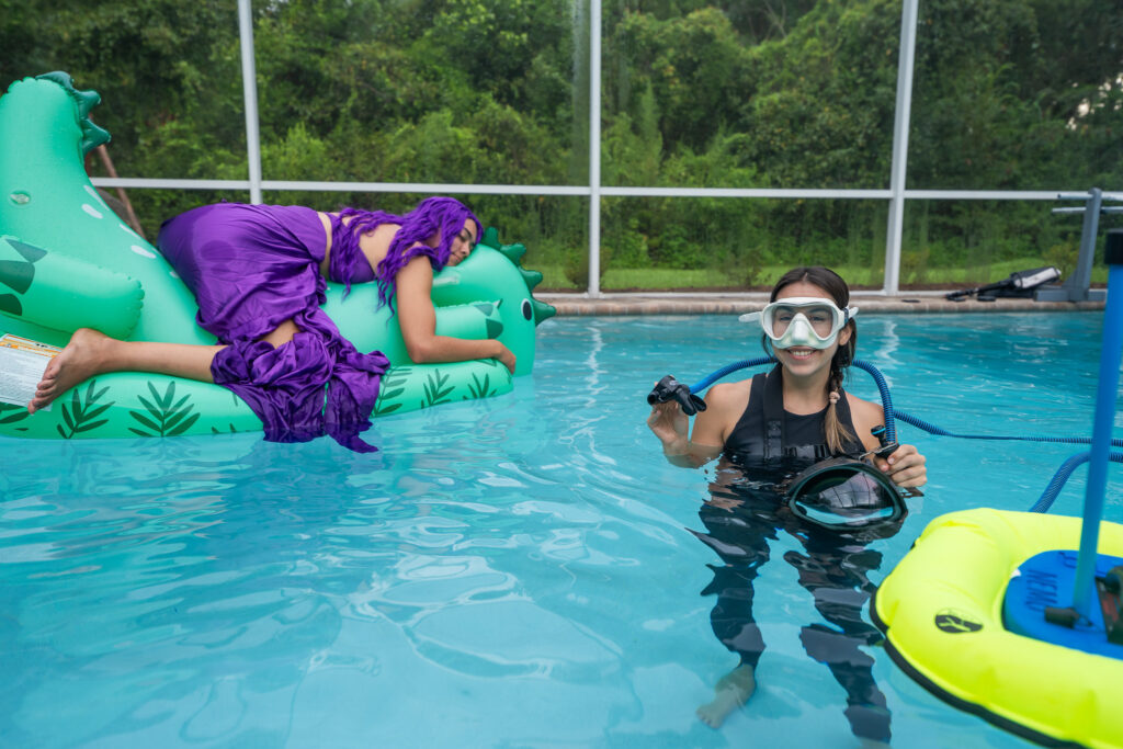 Photographer with BLU3 dive system in pool, preparing for underwater shoot, with model on an inflatable dinosaur behind her.