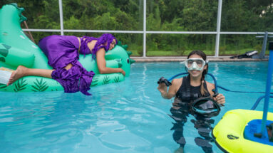 Photographer with BLU3 dive system in pool, preparing for underwater shoot, with model on an inflatable dinosaur behind her.