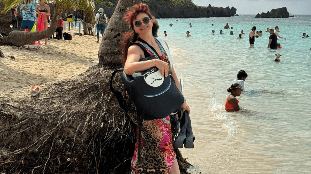 underwater-photographer-kate-ua-blu3-nomad-beach-dive Underwater photographer Kate UA standing on a tropical beach, holding a BLU3 Nomad dive system backpack, leaning against a palm tree.