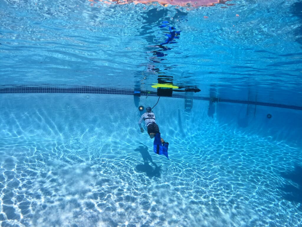Pool technician using a BLU3 Nomad tankless dive system to inspect the deep end of a commercial swimming pool for leaks. The diver is wearing fins and connected to the floating dive unit on the surface, demonstrating surface-supplied air operation in clear blue water.
