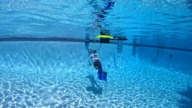 Pool technician using a BLU3 Nomad tankless dive system to inspect the deep end of a commercial swimming pool for leaks. The diver is wearing fins and connected to the floating dive unit on the surface, demonstrating surface-supplied air operation in clear blue water.