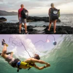 Two people with backpacks stand on rocky shore, while another swims underwater
