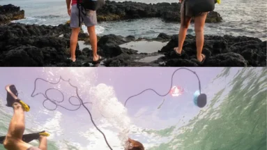 Two people with backpacks stand on rocky shore, while another swims underwater