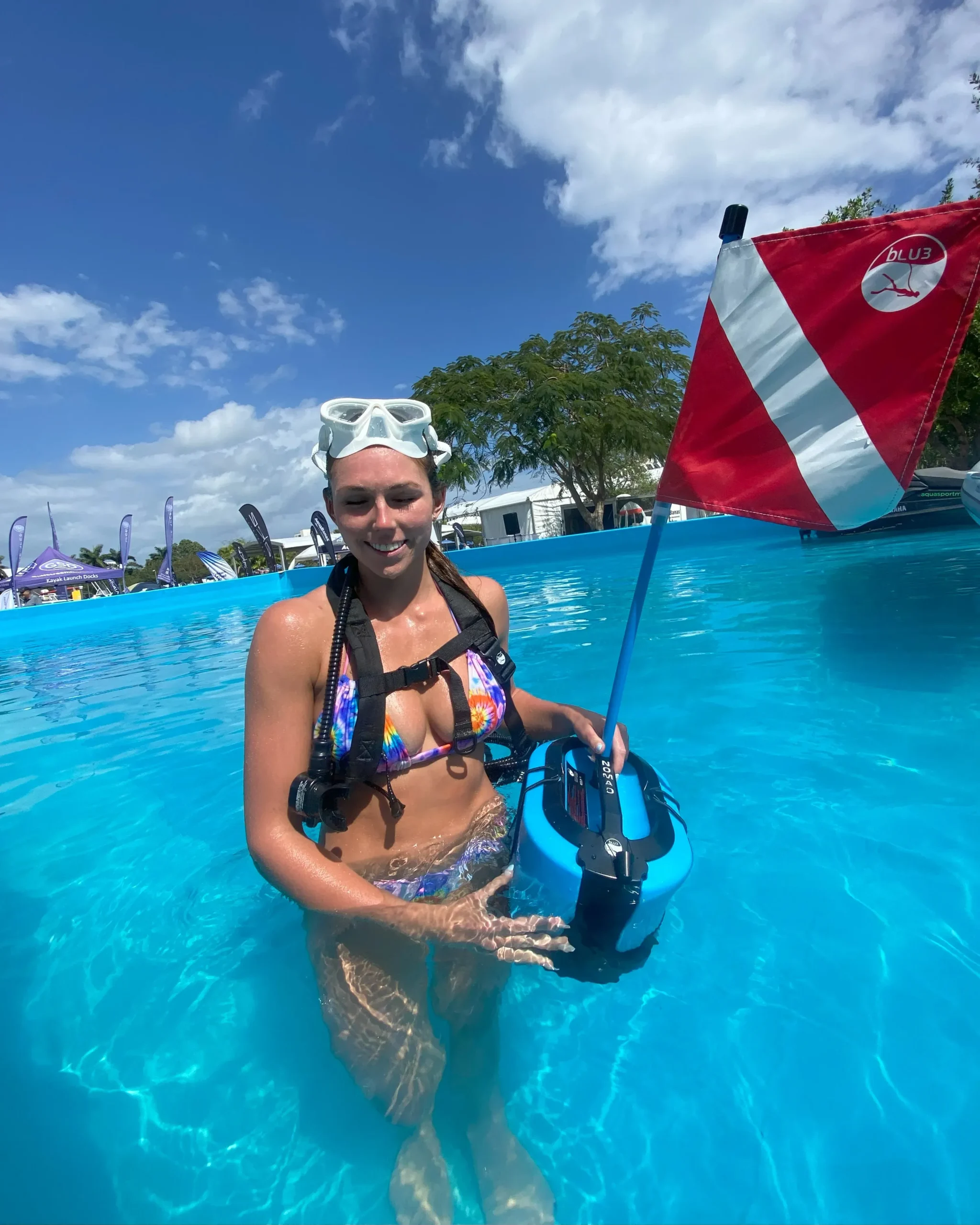 Woman in a colorful bikini holding a portable surface-supplied air system in a swimming pool, with a BLU3 flag in the background, demonstrating equipment for underwater inspections.